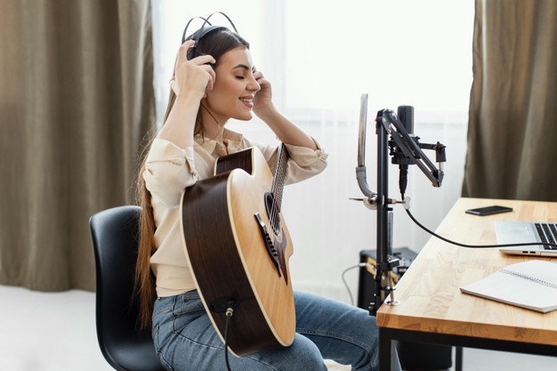 Side view of female musician putting on headphones to record song and play acoustic guitar 