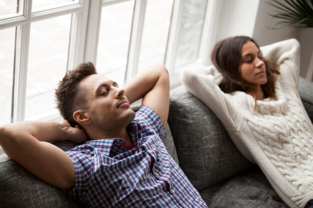 Young couple relaxing on comfortable sofa holding hands behind head 